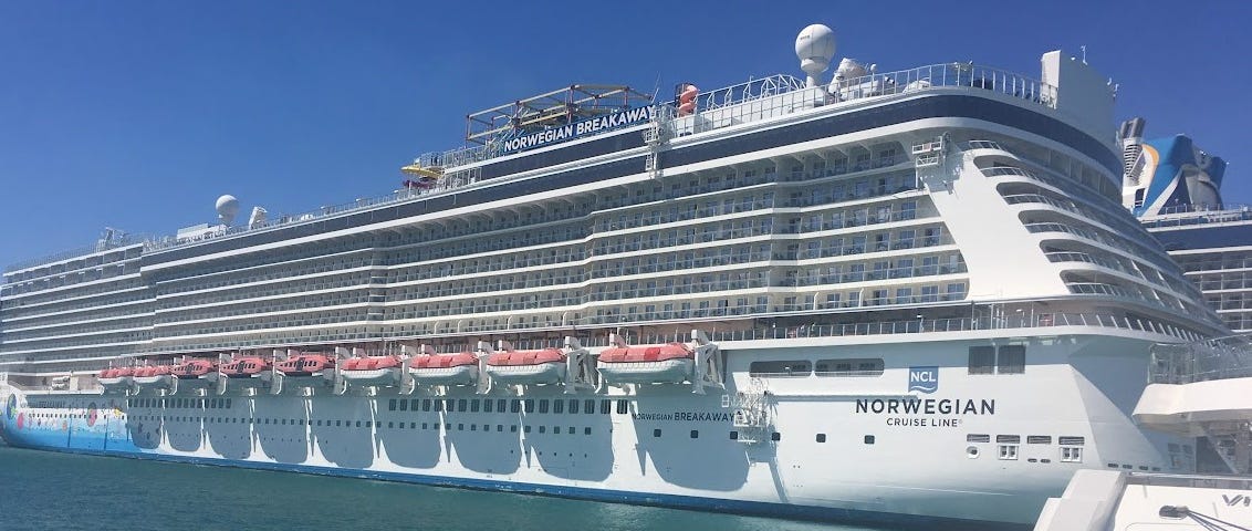 A large cruise ship sits docked near a pier. The words on the side of the ship announce its name, Norwegian Breakaway.