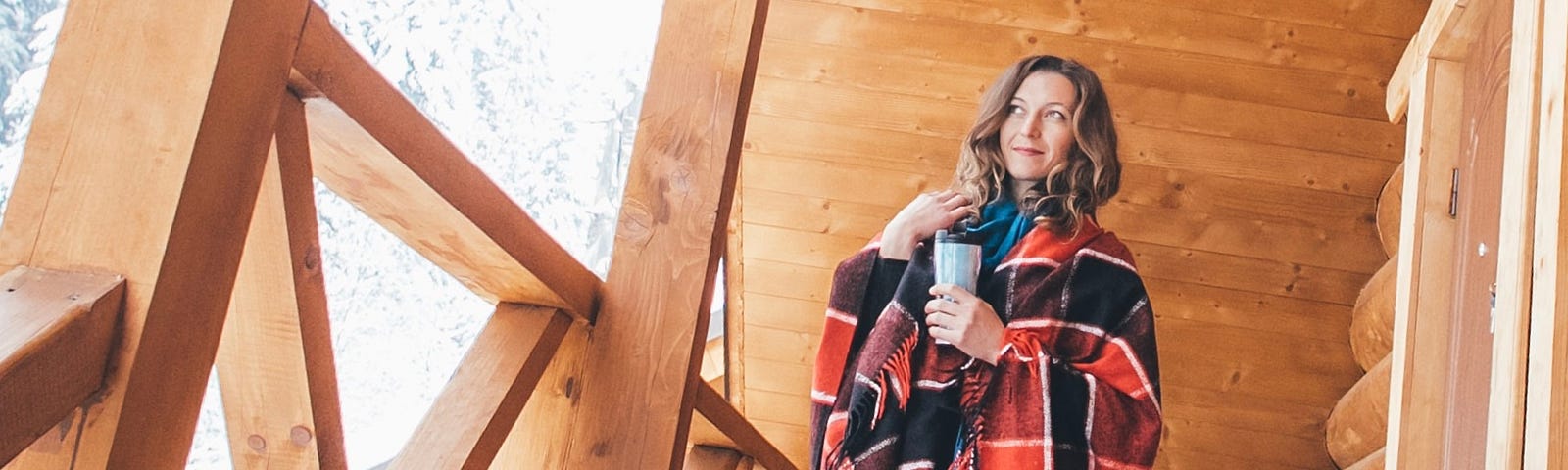 A woman in a wrap with a coffee in hand stands on the front porch of a log ski lodge in winter.