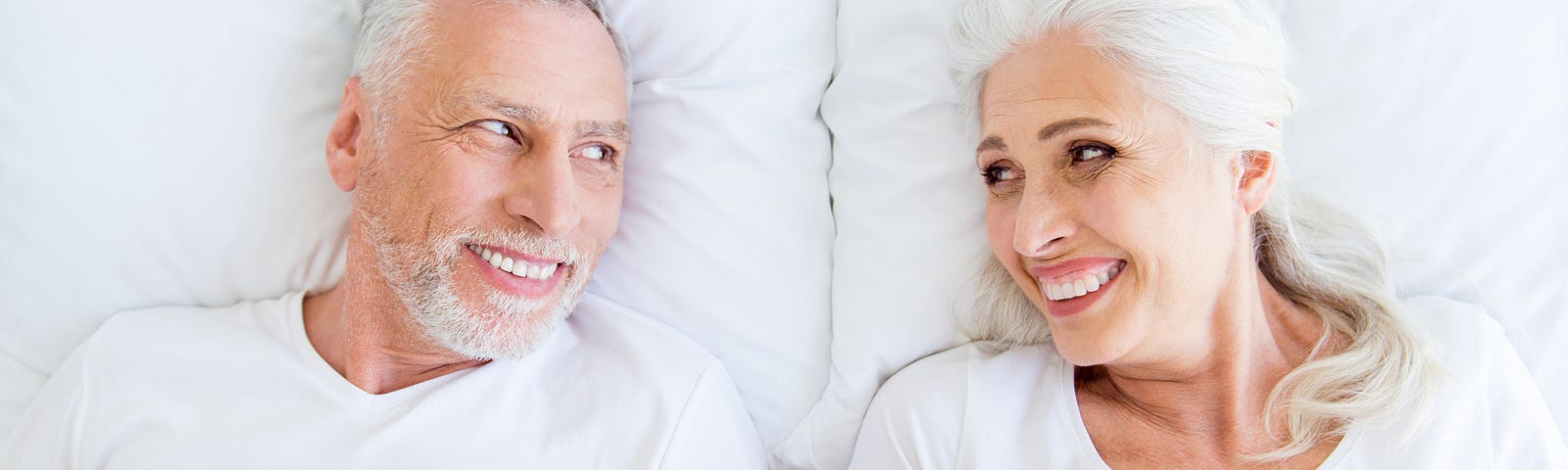 Older couple, lying in bed, smiling at each other.