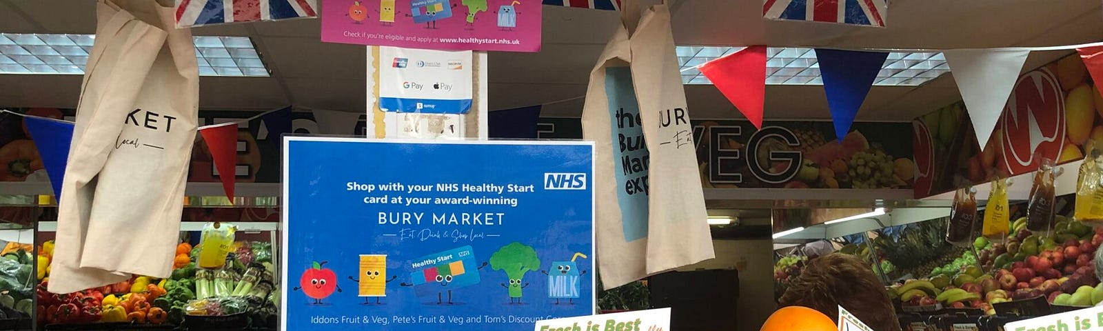 A fruit and veg stall at Bury Market with a sign saying “shop with your NHS Healthy Start card at your award-winning Bury Market”