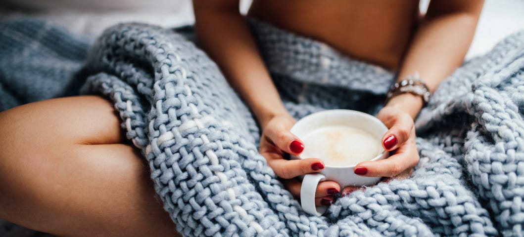 A blue chunky blanket is draped over the lap of a woman with red nails who holds a cup of coffee.