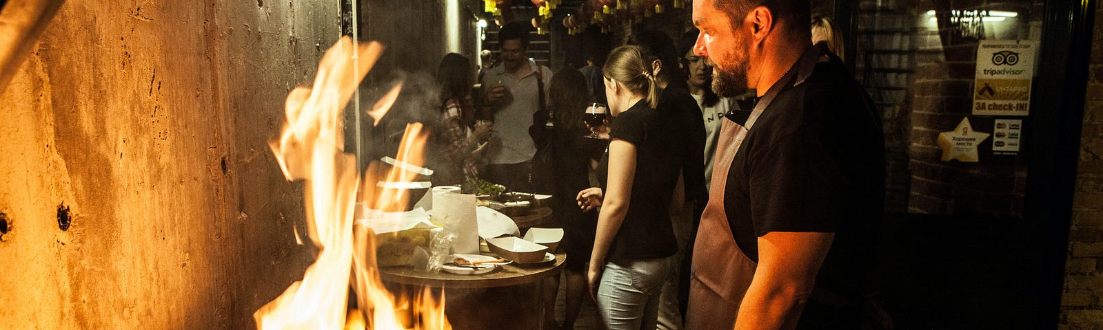 A man in a black shirt stands in front of a large flame. The background is a dimly lit, busy kitchen.