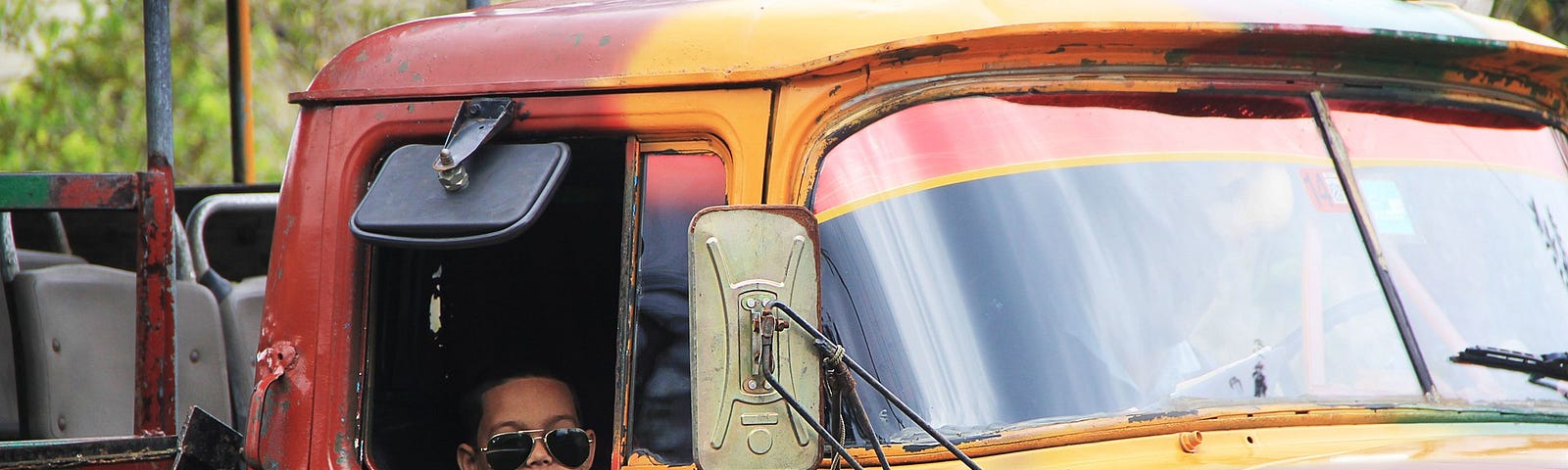 old red and yellow truck with small child wearing sunglasses sitting in the passenger seat, looking out the window.