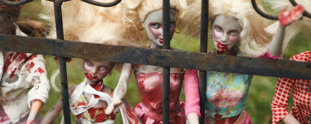 A group of Barbie dolls standing behind a fence. The dolls are painted and dressed to resemble zombies, with bloody hands and mouths.