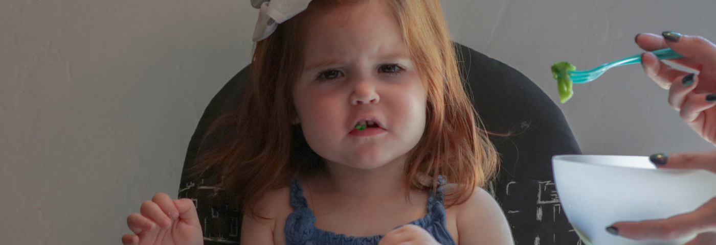 An adorable little red-headed girl clearly not happy eating broccoli in her high chair