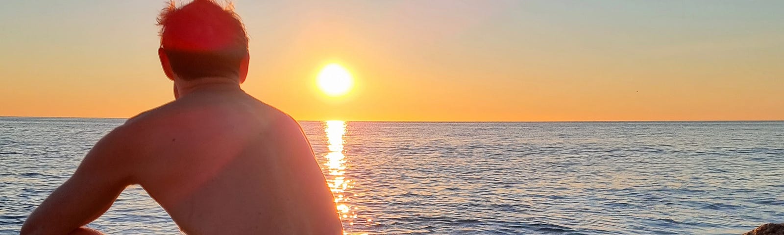 A man sitting on a rock on a beach in a swimming costume, watching the sunrise.