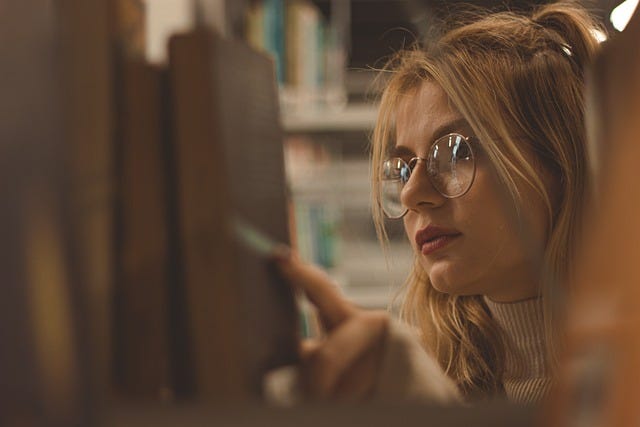 Young woman, blonde hair, glasses, books in foreground, library, shelf of books, novels, reading, selecting