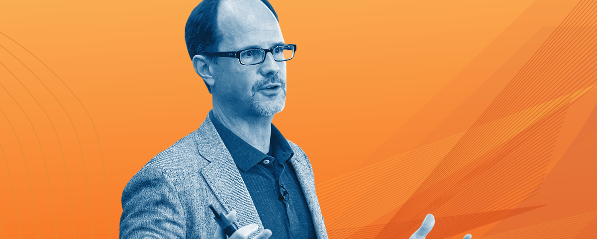 blue-scale photo of Rob Alexander wearing a blazer and gesturing mid sentence against an orange background.
