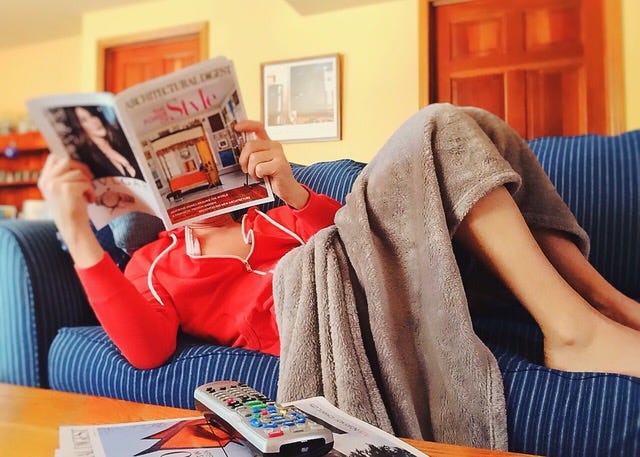 A woman lying on a blue couch covered in a blanket while reading a magazine.