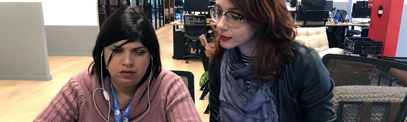 Camila e Iana estão sentadas na mesa de trabalho. Camila utiliza o celular com fones de ouvido enquanto Iana observa.