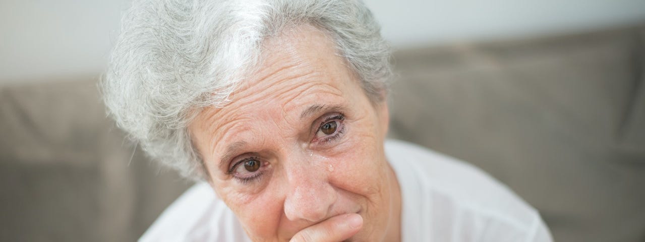 Photo of a woman with white hair, hand covering mouth, in tears, looking slightly away from the camera