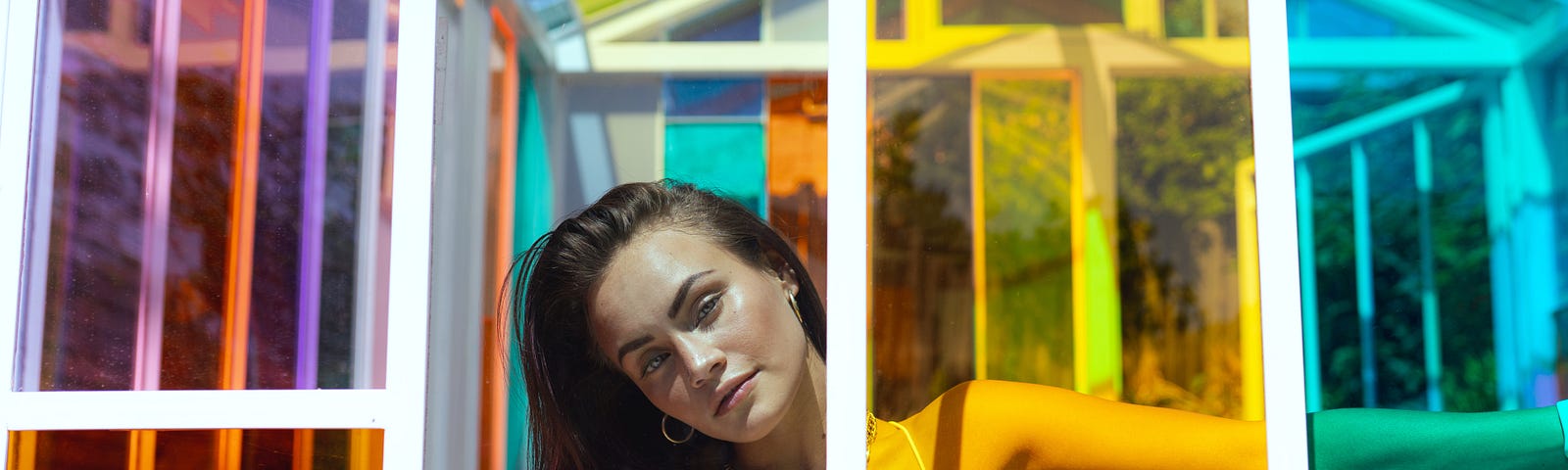 A woman with long dark hair in a white tank top casually looks through a gap in a house of colored panels