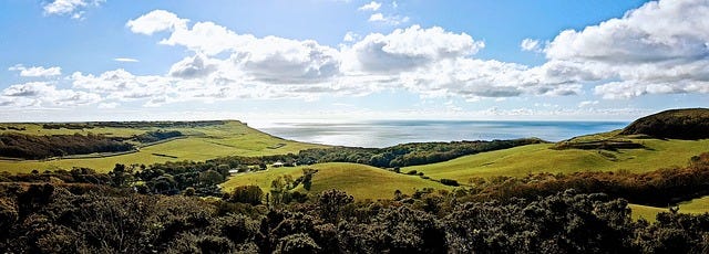 Sweeping green hills with trees in the forefront, the ocean at the back and a beautiful blue sky with white clouds