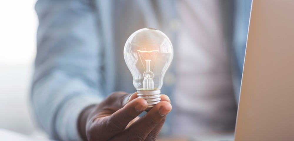 New idea concept. Unrecognizable black businessman holding illuminated light bulb in hand while sitting at workplace in office; ironically, Black inventor Louis Latimer contributed to the light bulb by inventing the carbon filament