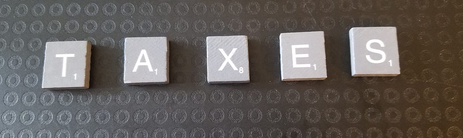 Grey wooden letter tiles on a black background. Letters spell TAXES.