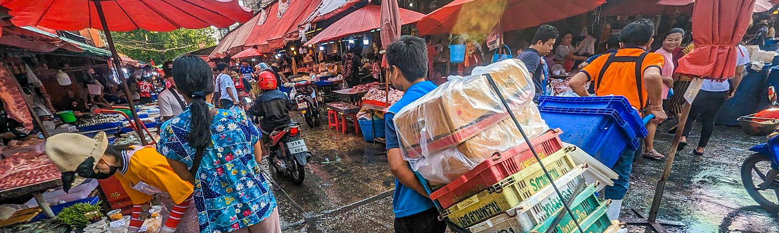 Man pulling pile of plastic bins behind of him as he walks through crowded market.
