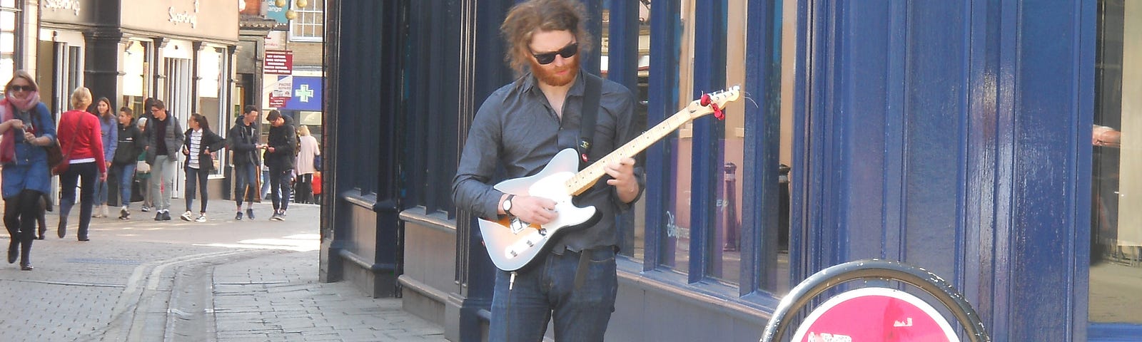 Man busking on a street corner in York, playing a Fender Telecaster electric guitar with his guitar case open at his feet to collect any coins donated to him by members of the public