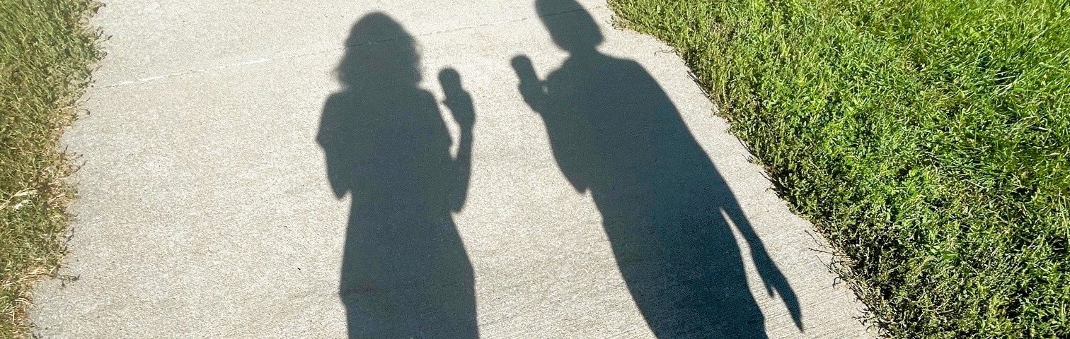 Shadows of two women — the author and her friend — as they walk together on a paved path edged by grass.