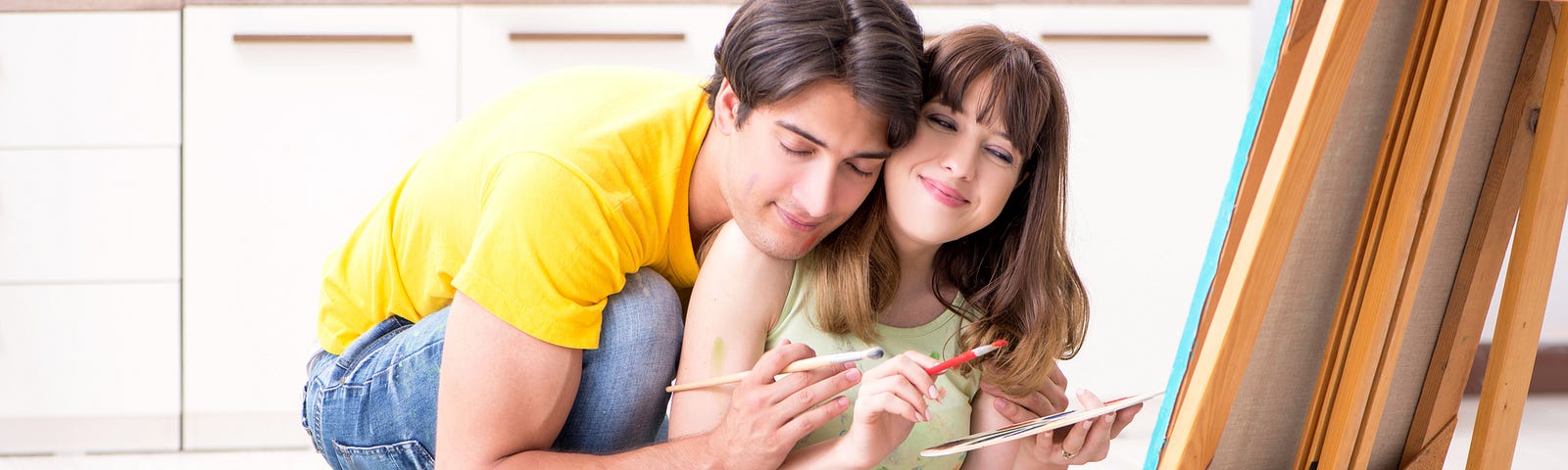 Man and woman embracing while holding paint brushes and sitting in front of a canvas (couples, relationships, painting, games, couple bonding)