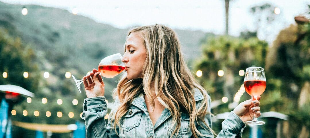 A woman sits on a table. She holds a wine glass containing a pink beverage in each hand and sips out of one of the glasses.