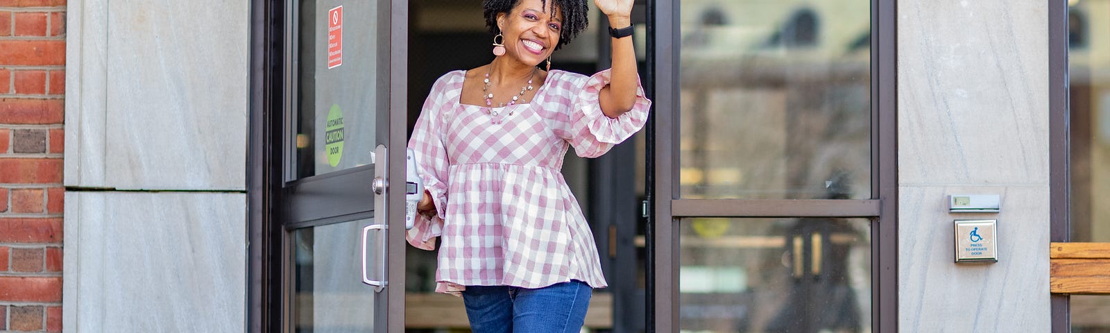 Tammy standing in an opened glass door waving, with a pink and white checkered shirt and jeans and grey boots.