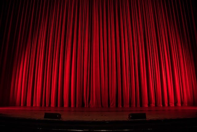 Theater stage, lit from below, with red curtain closed.