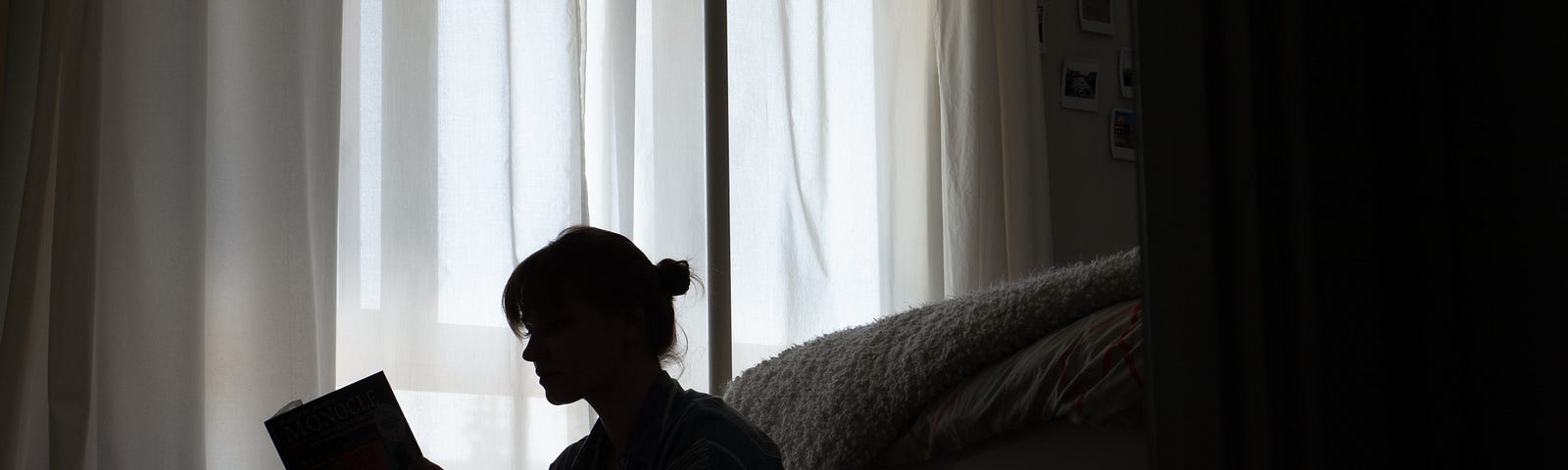 Dark image of a teenage girl reading on the floor next to a bed and curtained window
