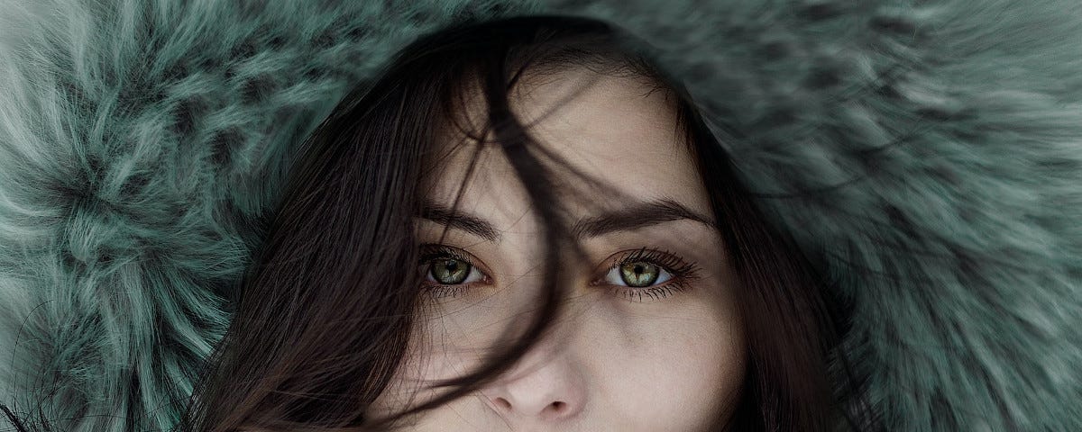 A portrait of a beautiful woman with long, straight, dark hair, full, red lips, and intense, grey-green eyes. A strand of her hair has fallen down her forehead, lying along the bridge of her nose before turning to her cheek. She is wearing a hood deeply lined with grey fur.