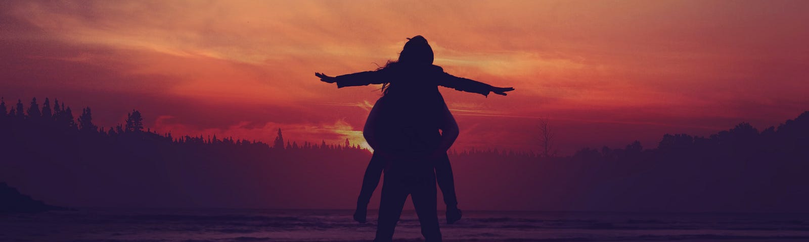 You can see the silhouette of a couple in front of the water’s edge. She’s on his shoulders with her arms out. The sun is setting with deep orange colours in the sky.