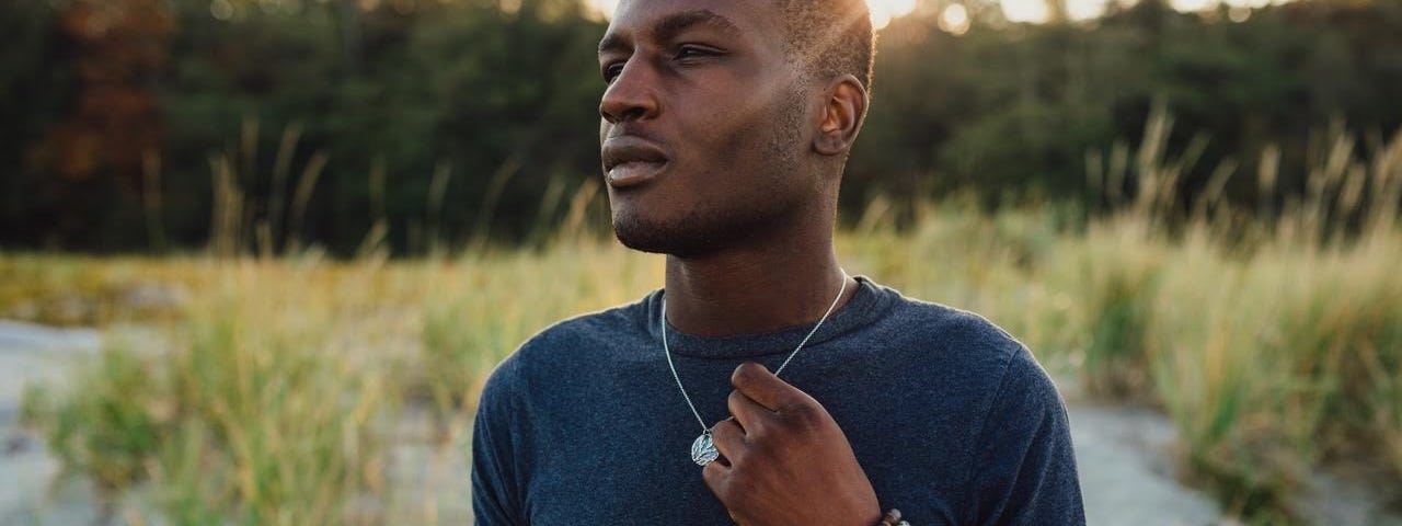 A Black man stands looking into the distance. He is wearing some bracelets and a necklace