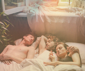 Two men lying in bed next to a woman, all cuddling/sleeping.