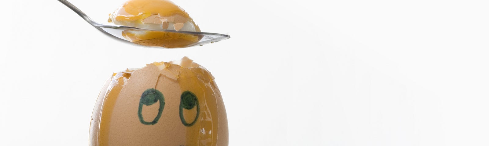 White background. Egg in egg cup. Egg with sad face drawn on it looking up. Egg is streaming with yoke from where a spoon above it has dug out the yoke.