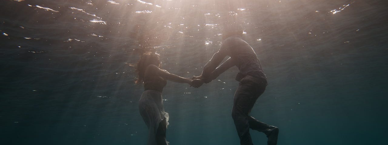 A fully-dressed couple holding hands under water, illuminated from above by sunlight.