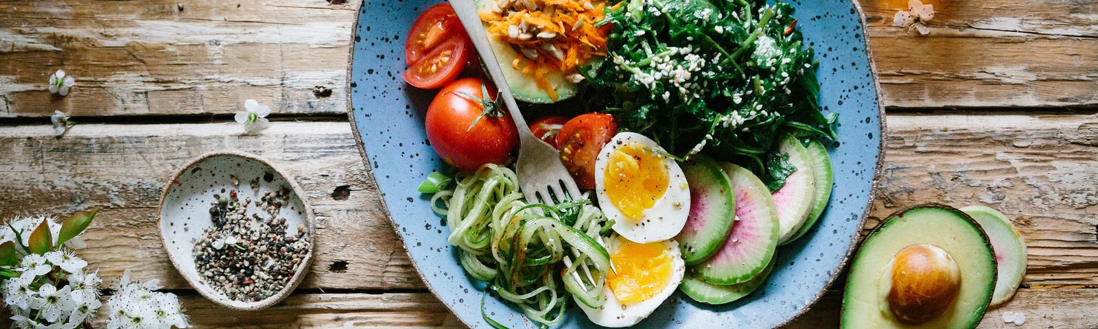 Photo of a Mediterranean style meal of greens, green whole grain pasta, sliced boiled eggs,  avocado, carrots and tomatoes on a blue plate. Glass of red wine on the side.