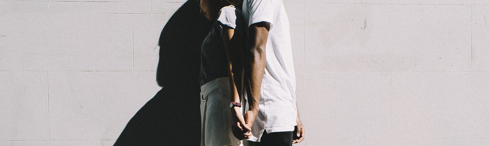 Man and woman standing back to back, holding hands, against a white wall with shadows.