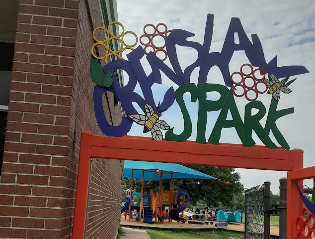 A colorful open gate topped with an artistic “Crenshaw Park” sign shows the shaded play structure and trees within the park.
