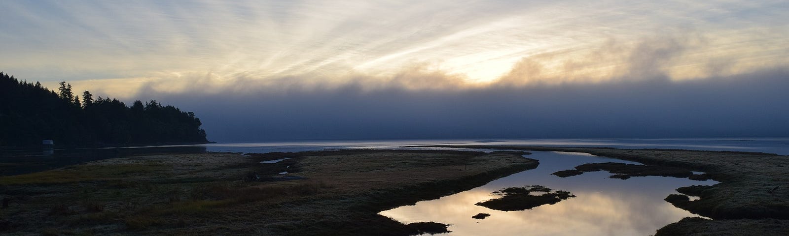 Photo of glassy tributary moving towards larger body/ocean at cloudy sunrise.