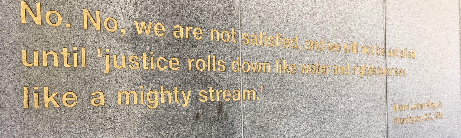 An inscription on a wall in San Francisco’s Dr. Martin Luther King, Jr. memorial reading, “No. No, we are not satisfied, and we will not be satisfied until ‘justice rolls down like water and righteousness like a mighty stream.’ Washington, D.C., 1963”.