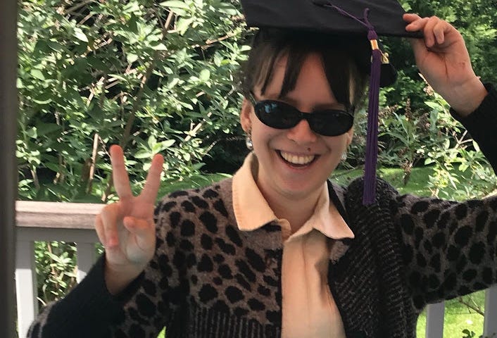 Girl with brown hair wearing a pink shirt, black patterened sweater, sunglasses, and a graduation cap, giving a peace sign.