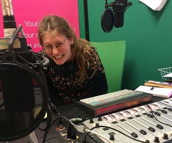 Photo of the poet Annie Muir sitting at an audio recording deck, with a joyful expression. The walls behind them are green.