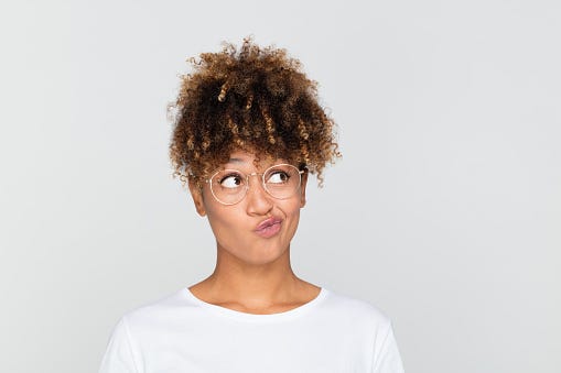 Mulher de pela negra, com cabelos crespos preso para cima, usando óculos de grau. Ela veste um camiseta branca e está com expressão de dúvida, o fundo onde a foto foi tirada é cinza.