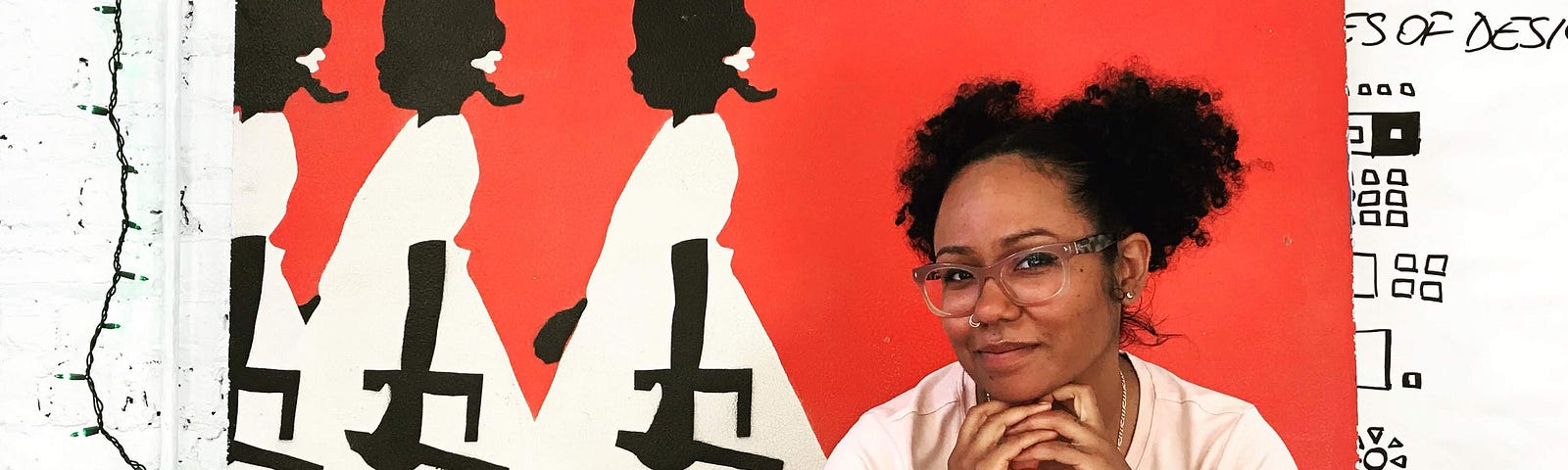 A smiling woman sits on the floor in front of a bright painting featuring three Black girls. Sharon has black hair & glasses