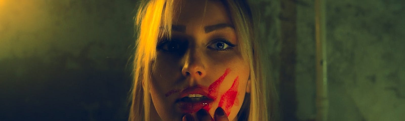 A young blond woman looks directly art the camera. Her hand is blood and she smears the blood into face.