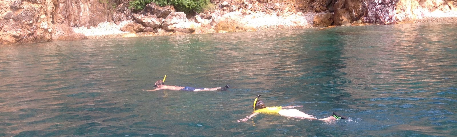 Two snorkelers float in calm water; wall of rock in the background.