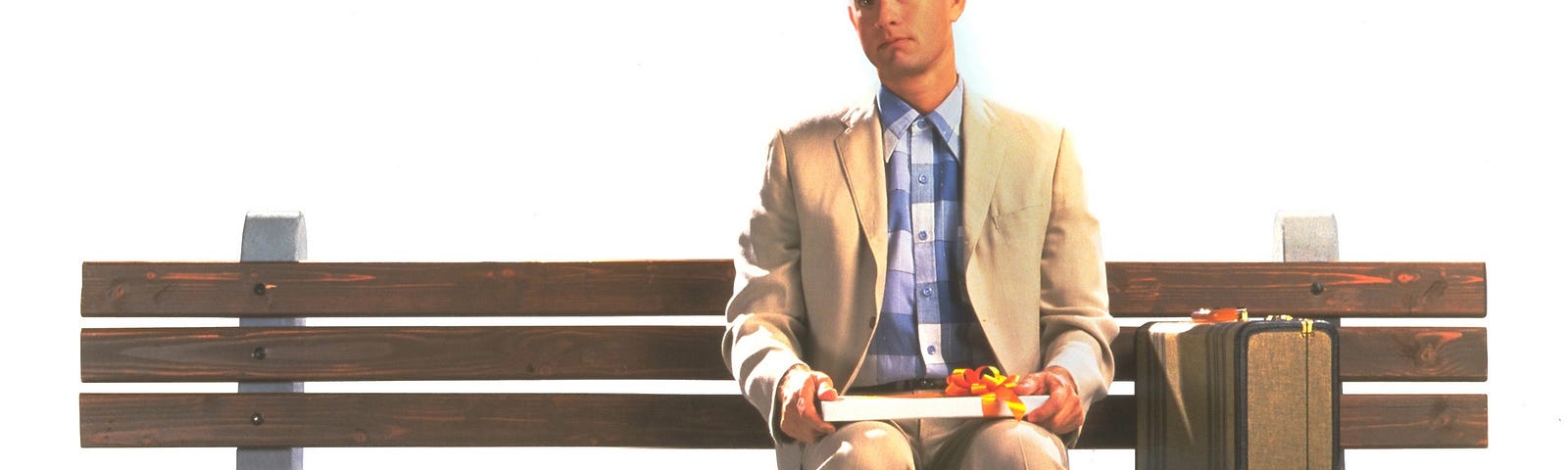 Scene from the movie Forrest Gump. Tom Hanks is sitting on a bench with a box of chocolates on his lap. A small suitcase is next to him on the bench. He is dressed in a tan suit with a blue plaid shirt.