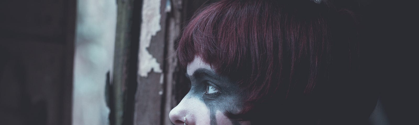 Woman at window with chipped brown paint behind her. She’s looking out window to the distance, her right arm is resting on the sill and her thumb is pressing on her bottom lip. She has black makeup painted in a splotch over her left eye and extending back beneath her maroon hair.