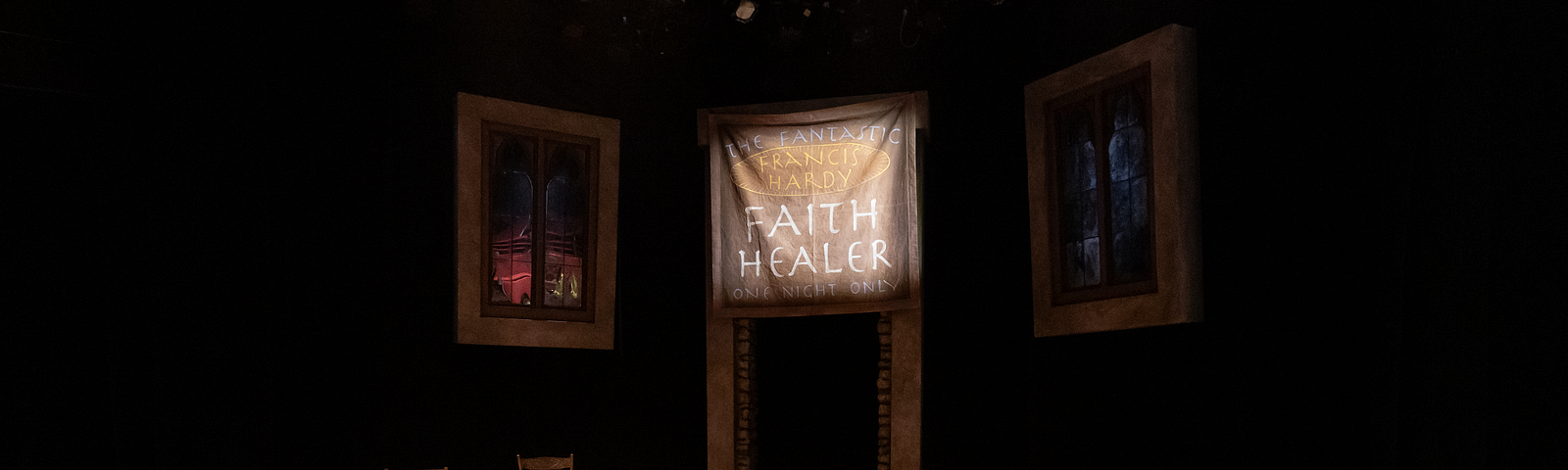 A theatrical set with wooden chairs, wooden floor, and a banner that reads “The Fantastic Francis Hardy, Faith Healer, One Night Only”