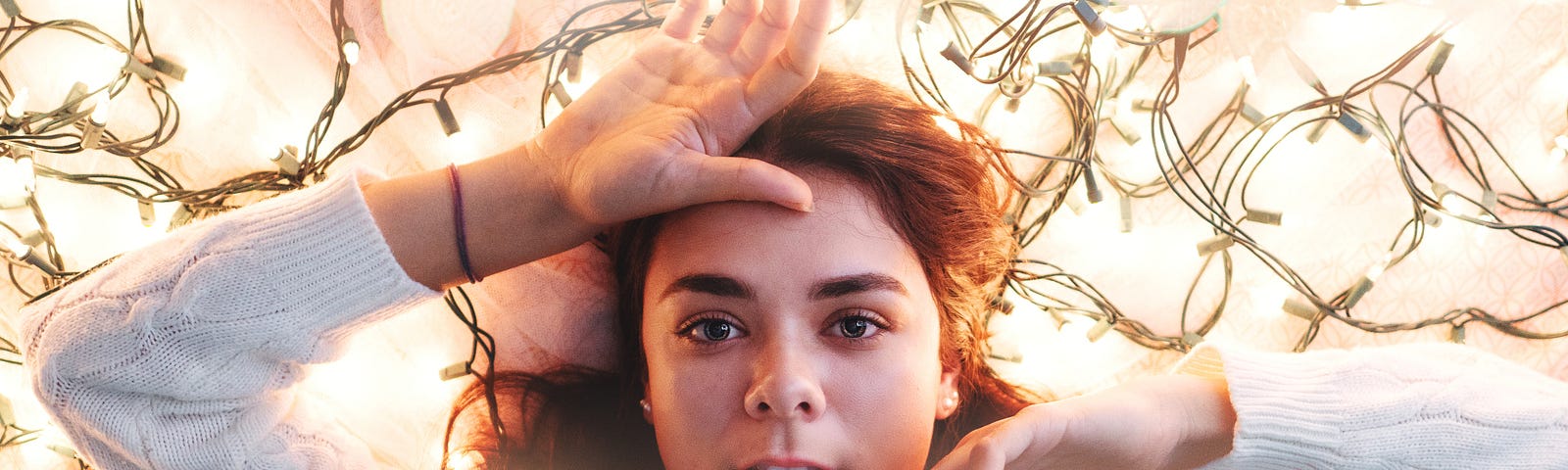 Woman with long dark hair wearing a white sweater lying in a pile of white Christmas lights. She is looking up at the camera.