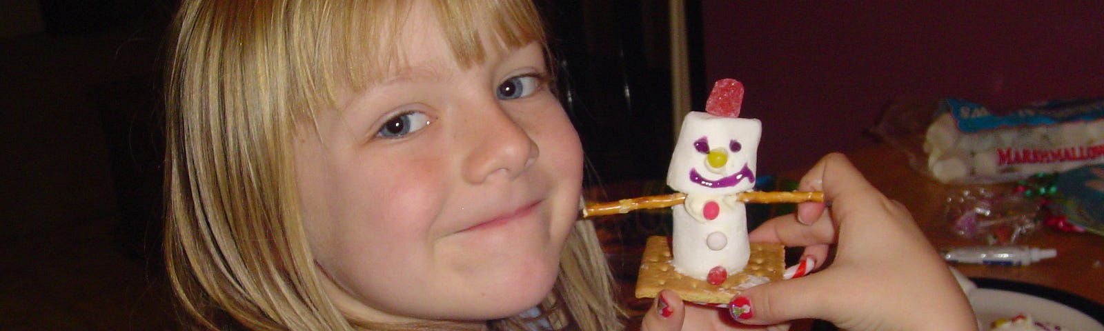 Young blonde girl holds two marshmallows on a graham cracker with gel frosting and straight toothpicks made to look like a snowman on a sled.