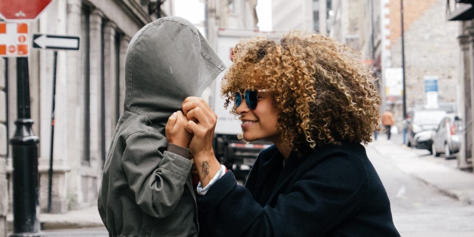 A mother crouches to smile at her young child on the street while they’re holding each other’s hands.